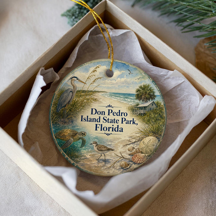 A round ornament with a beach scene and a bird is placed in a box with a white tissue paper lining.