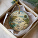 A round ornament with a beach scene and a bird is placed in a box with a white tissue paper lining.