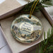 A decorative ornament in a box, featuring a scenic landscape with mountains, a cabin, and a wagon, along with the text "Donner Memorial State Park, California".