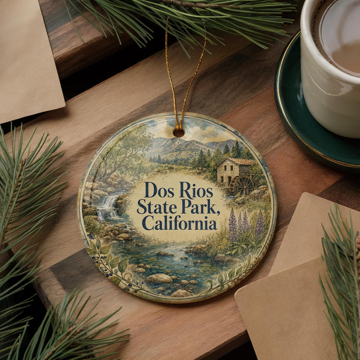 A decorative ornament featuring a scenic landscape with a watermill, surrounded by pine branches and a cup of coffee on a wooden surface.