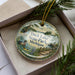 A decorative ornament in a box, featuring a scenic image of ducks in a lake surrounded by trees and a bridge.
