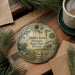 A decorative ornament featuring a rural scene with a windmill, a house, and a bird, surrounded by pine branches and flowers, placed on a wooden surface next to a cup of coffee.