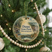 A round ornament hanging on a Christmas tree, featuring a scenic landscape with pine trees, a lake, and a bridge.