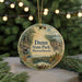 A circular ornament with a scenic landscape of a forest and a lake, featuring the text "Dunn State Park, Massachusetts" on it, is displayed in front of a blurred background with Christmas lights.