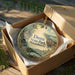 A decorative ornament in a box, featuring a scenic landscape with pine trees and a bridge, with the text "Dunn State Park, Massachusetts" printed on it.