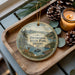 A decorative ornament featuring a scenic landscape of D Wroshak State Park, Idaho, with a deer, pine trees, and a lake. The ornament is displayed on a wooden tray, accompanied by pine cones and a lit candle.