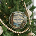 A round ornament hanging on a Christmas tree, featuring a scenic landscape with mountains, pine trees, and a lake.
