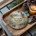 A decorative ornament with a beach scene and a lighthouse, placed on a wooden tray next to a lit candle and pine cones.