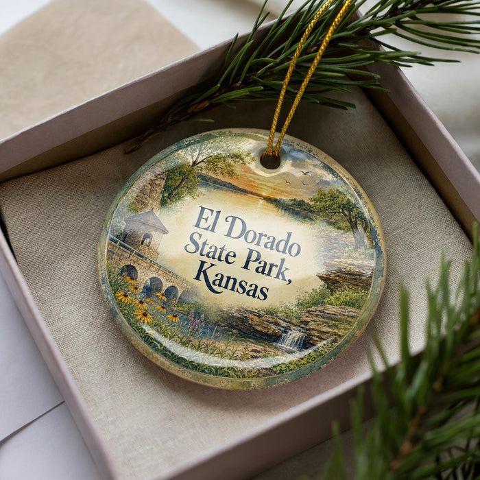 A decorative ornament in a box, featuring a scenic landscape with a bridge, trees, and a sunset.