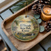 A decorative ornament with a scenic landscape of El Dorado State Park, Kansas, featuring a small house, trees, and a waterfall. The ornament is displayed on a wooden tray, with pine cones and a lit candle nearby.