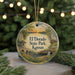 A round ornament with a painting of a scenic landscape, featuring a bridge, trees, and a sunset, hangs in front of a Christmas tree with lights.