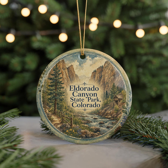 A round ornament with a scenic mountain landscape and a path leading through it, hanging in front of a blurred background with twinkling lights.