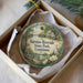 A round ornament with a painting of a park scene, featuring a house, trees, and a bird, is displayed in a box.