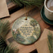 A round ornament with a tropical scene hangs on a wooden surface, surrounded by pine branches and a cup of coffee.