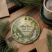 A round ornament with a scenic image of Falling Waters State Park in Florida, featuring a waterfall and lush greenery, is displayed on a wooden surface alongside a cup of coffee and some pine branches.