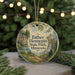 A round ornament with a scenic landscape of a waterfall and a house, surrounded by greenery, is displayed in front of a blurred background with twinkling lights.
