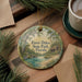 A round ornament with a painting of a bird and a cabin, surrounded by pine branches and a cup of coffee on a wooden surface.