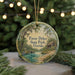 A round ornament with a painting of a bird and a cabin in a forest setting, surrounded by greenery and a body of water.