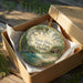 A round ornament in a brown box, featuring a scenic landscape with a lake, trees, and a cabin, with the text "Faver-Dykes State Park, Florida" printed on it.