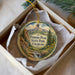 A round ornament with a scenic mountain landscape and text, placed in a box with a white tissue paper lining.