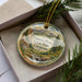 A decorative ornament in a box, featuring a scenic landscape with mountains, pine trees, and a waterfall.