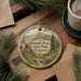 A round ornament with a scenic mountain landscape and text, hanging on a wooden surface next to a cup of coffee and pine branches.