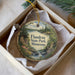 A decorative ornament in a box, featuring a scenic landscape with a lake, trees, and a cabin.