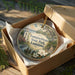 A decorative ornament in a box, featuring a scenic landscape with a building, trees, and flowers.