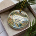 A decorative ornament in a box, featuring a scenic landscape with mountains, trees, and wildlife.