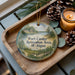 A round ornament with a scenic landscape of Fort Custer Recreation Area, Michigan, is displayed on a wooden tray alongside pine cones and a lit candle.