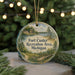 A round ornament with a painting of a scenic landscape, featuring a lake, forest, and a building, hangs in front of a Christmas tree with twinkling lights.