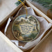 A round ornament with a painting of Fort King George Historic Site, featuring a house, a boat, and a tree, is displayed in a box.