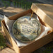 A round ornament in a box, featuring a scenic image of Fort King George Historic Site in Georgia.