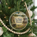A round ornament hanging on a Christmas tree, featuring a scenic landscape with a castle, a river, and a field with purple flowers.