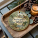 A decorative ornament with a painting of Fort Washington State Park, Maryland, sits on a wooden tray alongside pine cones and a lit candle.