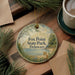 A round ornament with a scenic image of a fox in a park, surrounded by pine branches and a body of water, is displayed on a wooden surface alongside a cup of coffee and some papers.