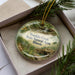 A round ornament with a scenic image of Franklin Creek, a natural landmark in Illinois, displayed in a box with pine branches.