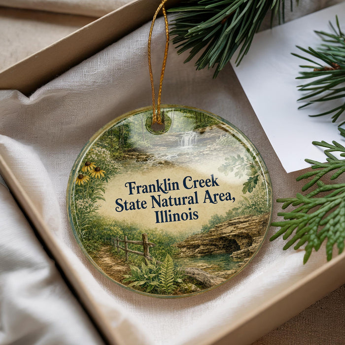 A round ornament with a scenic landscape of Franklin Creek, the State Natural Area in Illinois, displayed in a box with pine branches and a white card.
