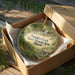 A round ornament in a brown box, featuring a scenic image of Franklin Creek in Illinois.