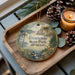 A wooden tray with a decorative ornament featuring a scenic landscape of Frontenac State Park, Minnesota, along with pine cones and a lit candle.