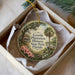 A round ornament with a painting of a house and palm trees, hanging from a string inside a box.