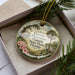 A decorative ornament in a box, featuring a scenic landscape with palm trees, flowers, and a house, with the text "Gamble Plantation Historic State Park, Florida" printed on it.