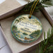 A decorative ornament in a box, featuring a scenic landscape with a lake, pine trees, and a boat, along with the text "Garden Island State Recreation Area, Minnesota".