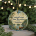 A circular ornament with a scenic landscape featuring a lake, pine trees, and a canoe, hanging in front of a blurred Christmas tree with lights.