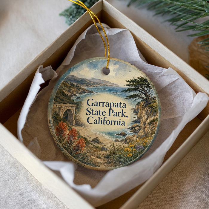 A decorative ornament in a white box, featuring a scenic landscape with a bridge, trees, and a body of water.