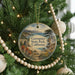 A round ornament hanging on a Christmas tree, featuring a scenic landscape with a bridge and mountains in the background.