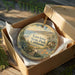 A decorative ornament in a brown box, featuring a scenic landscape with mountains, a house, and a body of water.