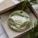 A decorative ornament in a box, featuring a scenic landscape with a river, cabin, and mountains.