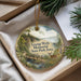 A decorative ornament with a scenic landscape featuring a bridge, deer, and trees, placed in a box with pine branches.