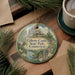 A decorative ornament featuring a castle surrounded by trees and a river, with the text "Gillett Castle State Park, Connecticut" displayed on it. The ornament is placed on a wooden surface, with a cup of coffee and some papers nearby.